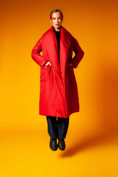 Trendy female in oversize coat jumping against yellow background — Stock Photo, Image