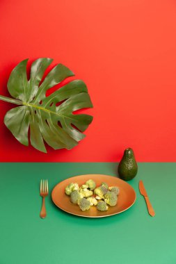 Taze çiğ brokoli tabağı ve çatal bıçak takımı Monstera yaprağı yakınındaki kırmızı masaya yerleştirilmiş.