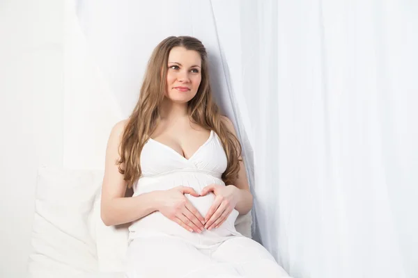 Piękna kobieta w ciąży w białej sukni w pobliżu okna — Zdjęcie stockowe