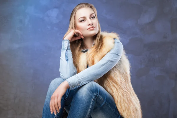 Elegant woman sits wearing blue jeans and fur vest — Stock Photo, Image