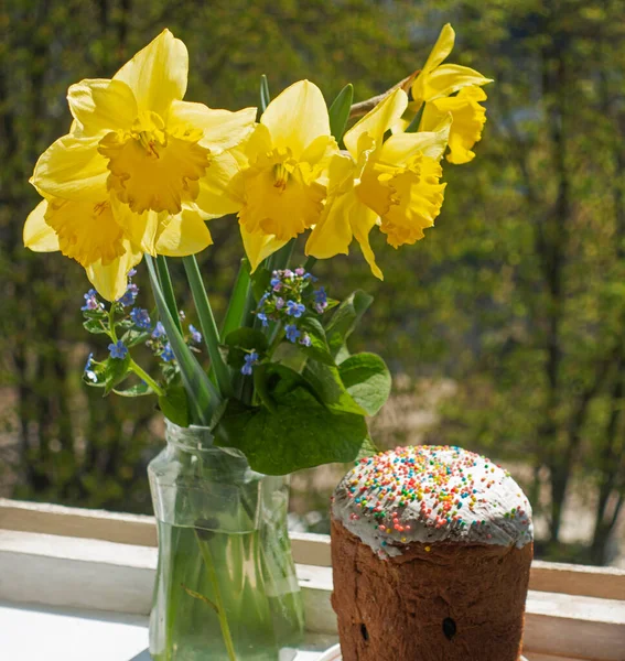 Spring bouquet of yellow daffodils and Easter cake by the window against the background of trees