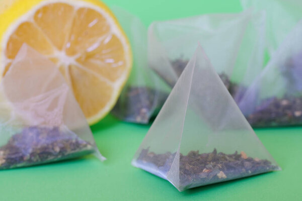 На зеленой поверхности рядом с нарезанным лимоном лежит пирамидальный чайный пакетик со вкусом зеленого чая с ломтиками лимона и листьями мяты. Макро. Мелкая глубина резкости