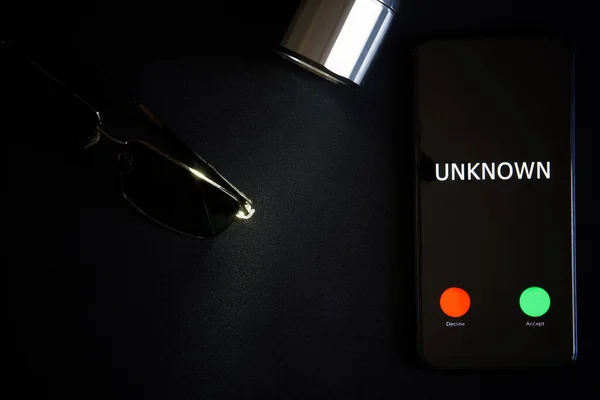 A smartphone with an unknown call lies on a black surface next to sunglasses and a flashlight. The problem of anonymity and espionage with modern gadgets. Flatlay