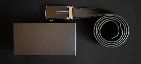 ブラックレザーのメンズベルトで ゴールドトーンの自動バックルがギフトボックスの横にある暗い背景にあります 接近中だ バナー — ストック写真