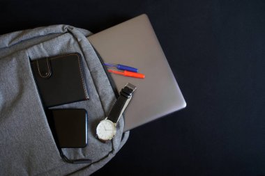 Laptop ya da ultraviyole çantalı küçük gri bir tekstil sırt çantası, bir cüzdan, bir akıllı telefon ve bir kol saati siyah arka planda yer alır. Şehir gezgini, programcı ya da öğrencinin özellikleri. Yakın plan.