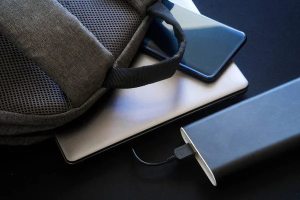 A large black external battery - power bank lies on the table next to a gray textile backpack, laptop or ultrabook and smartphone. Travel and travel with gadgets. Close-up
