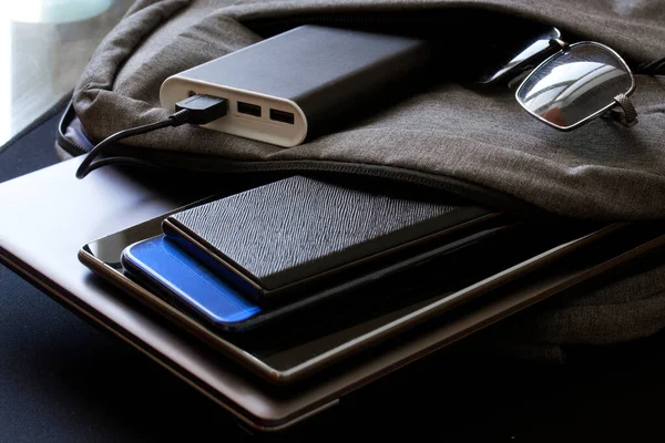 İçinde bir sürü alet ve harici batarya bulunan bir çanta ya da çanta siyah bir yüzey üzerinde duruyor. İki akıllı telefon, dizüstü bilgisayar veya ultrabook, güneş gözlüğü ve tablet. Modern inek seyahat kiti. Yakın plan.