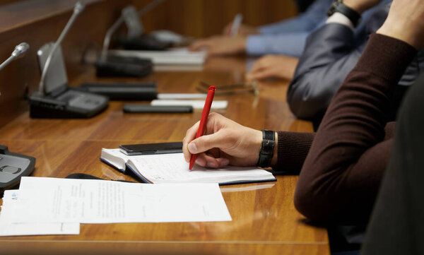 A man writes something in a diary notebook during a business meeting, lecture in college, or negotiations. Table with microphones and speakerphones. No face