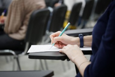 Bir kız bir dikte yazar ya da izleyiciler arasında doküman doldurur, elinde yazı standı olan bir okul sandalyesine oturur. Yakın plan. Yüzü yok