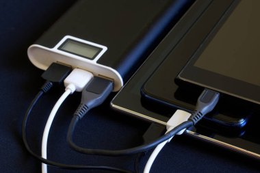 Three USB gadgets - two smartphones and a tablet are charged from one external battery - powerbank. Dark background. The concept of recharging a variety of smart devices. Close-up clipart
