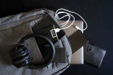 Birçok modern cihaz: bir dizüstü bilgisayar, tablet, akıllı telefonlar ve deri cüzdan harici bir bataryayla birlikte gri bir sırt çantasında. Seyahat ederken ve seyahat ederken aletleri şarj etme kavramı. Yakın plan.