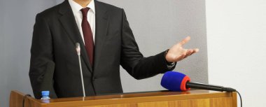 Politikacı, öğretmen ya da avukat kürsüden konuşuyor. Jest ve mikrofon. Yüzü yok