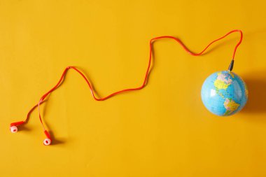 Red wired earbuds are connected to a small globe. The concept of broadcasting, webcasting, podcasting and broadcasting worldwide popular audio hits. Yellow background