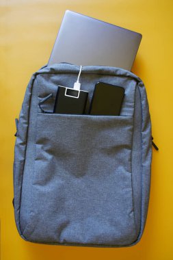 Gray textile city backpack containing silver laptop, smartphone and an external battery - power bank on a yellow background. Study, travel and trip with gadgets. Yellow gradient. View from above