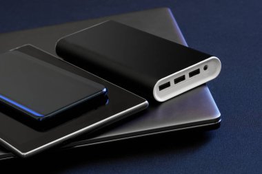 An external battery is a power bank along with gadgets: a smartphone, a thin laptop and a tablet. The concept of recharging mobile devices during work, business or travel. Black background