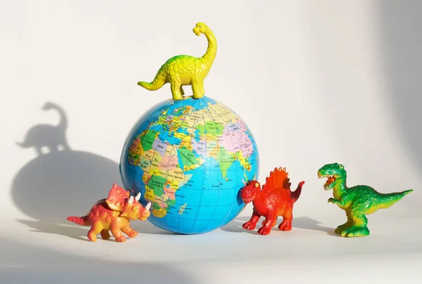 Funny toy dinosaurs are standing around a globe - the globe. The concept of paleontology and the study of dinosaurs in school and college. White background. Close-up