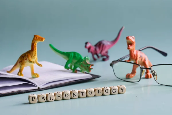 Paleontology inscription next to toy dinosaurs, open book or textbook and glasses. The concept of studying paleontology and dinosaurs in school, university and college. Pastel background