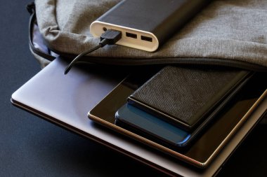 Modern mobil cihazlar, iki akıllı telefon ve dizüstü bilgisayar bir güç bankasının yanında gri bir sırt çantasında yatıyor. Seyahat ederken aletlerinizi şarj edin. Yakın plan.
