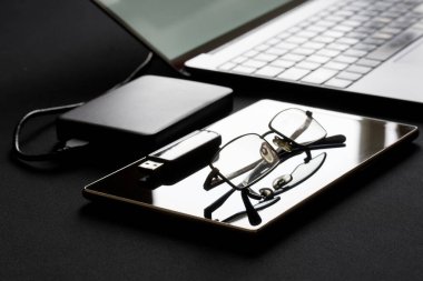 Açık ince bir dizüstü bilgisayar tabletin, harici hard diskin, USB belleğin ve gözlüklerin yanında durur. Arka ışıklandırma. Veri güvenliği, işyeri, uzaktan kumanda ve ofis işi kavramı. Yakın plan.