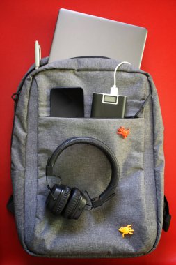 İçinde cihazlar olan modern laptop çantaları: kulaklıklar, akıllı telefon, dizüstü bilgisayar ve dış batarya. Kırmızı arka plan. Çalışırken, seyahat ederken ve seyahat ederken aletleri şarj etme kavramı. Yakın plan.