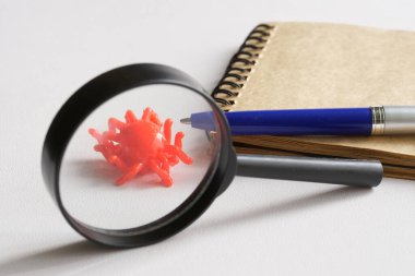 Oyuncak, bir defter ve kalemin yanındaki büyüteç altındaki bir virüs. Virüs mutasyon çalışması ve araştırma konsepti. Yeni bulaşıcı türlerin analizi. Beyaz arka plan. Yakın plan.