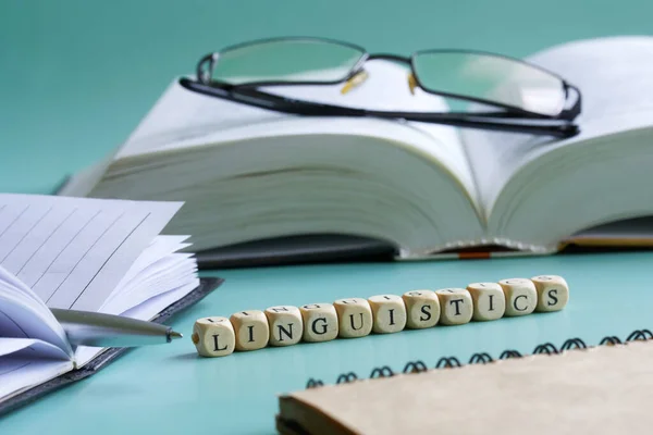 The word linguistics next to notepad, pen, open book and glasses. Concept for the study of liberal arts and linguistics at college and university. Pastel background. Selective focusing. Macro