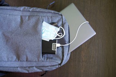 Gri tekstil şehir sırt çantası, içinde gümüş bir dizüstü bilgisayar, güç bankası, tablet ve ahşap bir masanın üzerinde koruyucu bir tıbbi maske var. Bir salgın sırasında aletlerle çalışmak, seyahat etmek ve gezmek. Yakın plan.