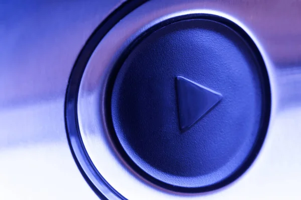 Round embossed play button on a light polished metal surface of a modern gadget. Blue neon lighting for a nightclub or disco. Macro. Close-up