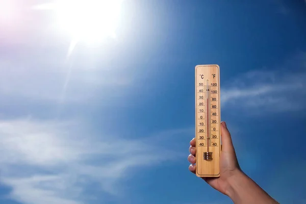 A thermometer with red scale in hand on the sky and sun background. Hot weather concept