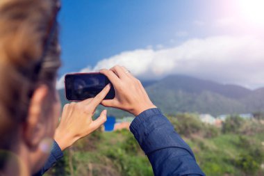 Kısa sarı saçlı, akıllı telefon kullanan kadın dağ manzaralı bir fotoğraf çek.