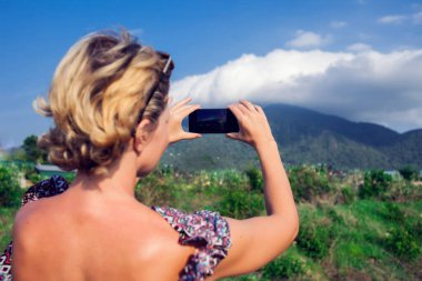 Kısa sarı saçlı, akıllı telefon kullanan kadın dağ manzaralı bir fotoğraf çek.