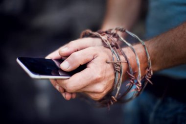 Smartphonein hands with barbed wire on a dark background. Concept of Social Disease , mobile phone addicted. clipart