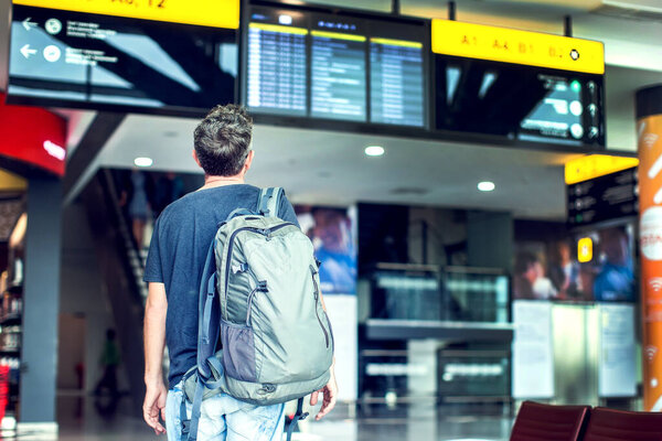 A young male traveler with a backpack in casual style looks at the information board at the airport. Getting information about the flight.