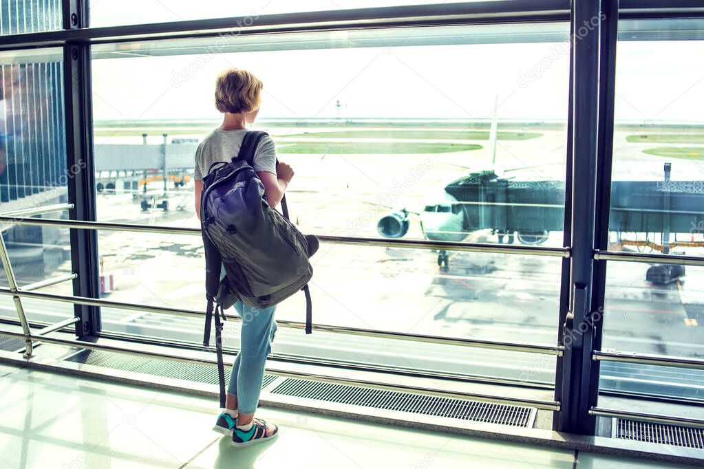 Travel tourist standing with luggage watching at airport window. Unrecognizable woman with short hair looking at lounge looking at airplanes at boarding gate before departure. Travel lifestyle.