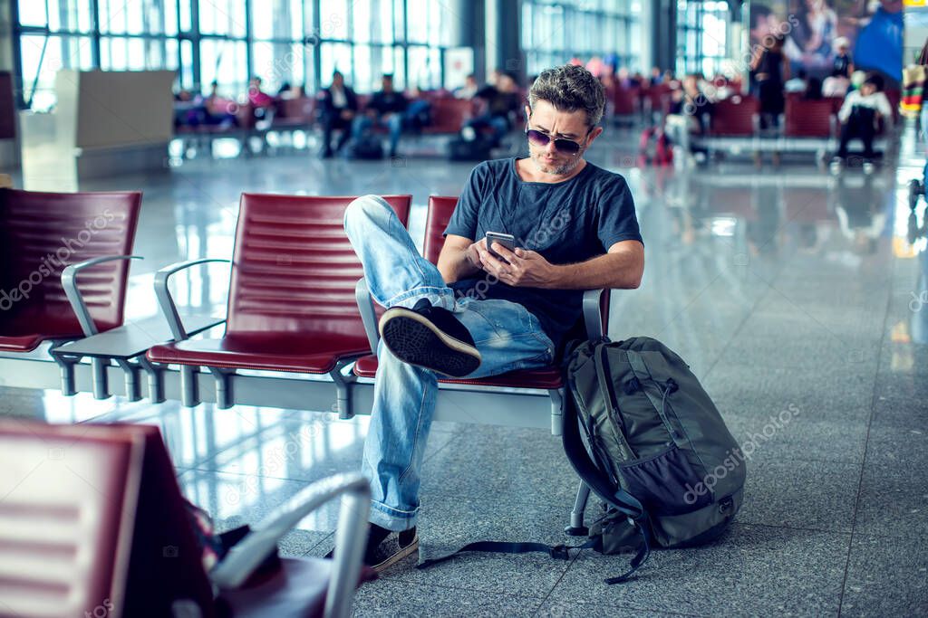 Young man checking his phone while waiting his flight in the airport at the departure area