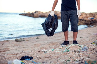 Siyah gömlekli, şortlu, beyaz eldivenli ve büyük siyah paketli bir adam sahilde çöp topluyor. Çevre koruma ve gezegen kirliliği kavramı