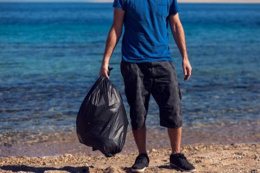 Siyah çantalı gönüllü bir adam sahilde çöp topluyor. Çevre kirliliği kavramı