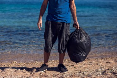 Siyah çantalı gönüllü bir adam sahilde çöp topluyor. Çevre kirliliği kavramı