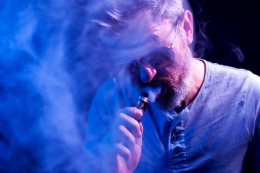 Güneş gözlüklü sakallı adam mavi arka planda sigara içiyor. Sigara bağımlılığı konsepti