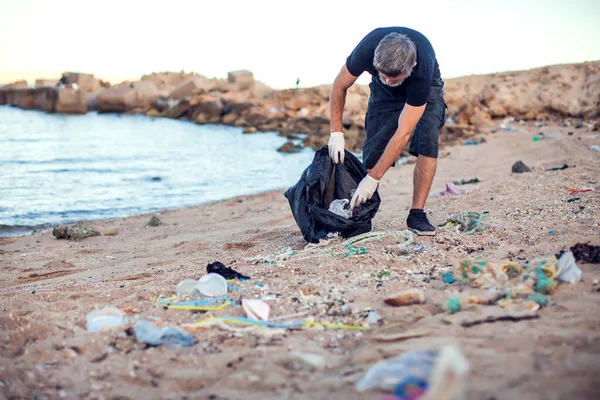白い手袋とビーチでゴミを収集する大きな黒いパッケージと暗いシャツやショートパンツの男 環境保護と地球汚染の概念 — ストック写真
