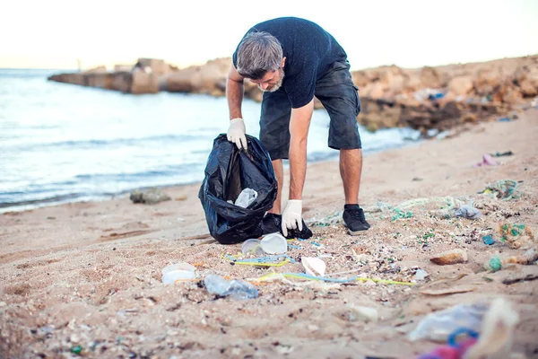 셔츠와 반바지를 장갑을 커다란 꾸러미를 남자가 해변에서 쓰레기를 수거하고 — 스톡 사진