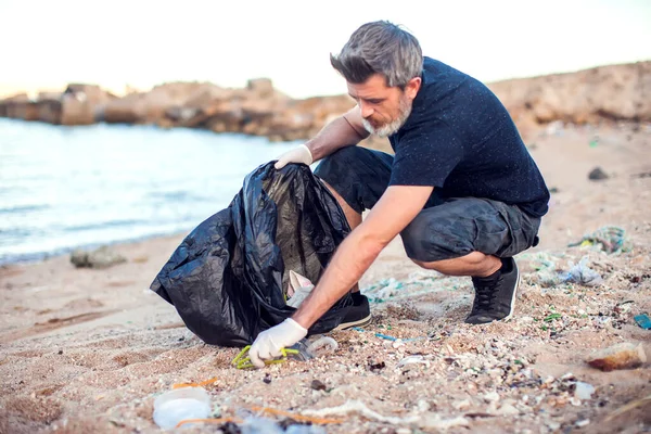 白い手袋とビーチでゴミを収集する大きな黒いパッケージと暗いシャツやショートパンツの男 環境保護と地球汚染の概念 — ストック写真