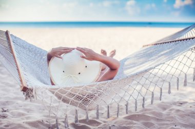 Hamakta dinlenen beyaz şapkalı bir kadın tatilde güneşleniyor.