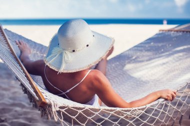 Hamakta dinlenen beyaz şapkalı bir kadın tatilde güneşleniyor.