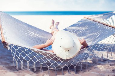 Hamakta dinlenen beyaz şapkalı bir kadın tatilde güneşleniyor.