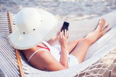 Tatil ve teknoloji. Şapkalı ve mayolu genç bir kadın kumsalda hamakta yatarken akıllı telefon kullanıyor..