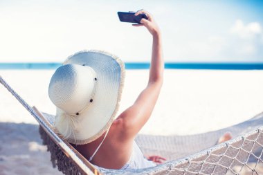 Tatil ve teknoloji. Şapkalı ve mayolu genç bir kadın kumsalda hamakta yatarken akıllı telefon kullanıyor..