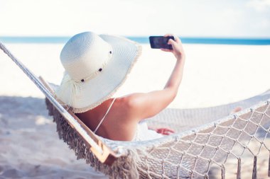 Tatil ve teknoloji. Şapkalı ve mayolu genç bir kadın kumsalda hamakta yatarken akıllı telefon kullanıyor..