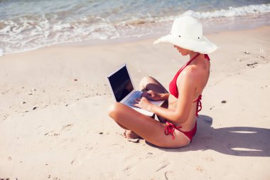 Gülümseyen kız bikini ve hasır şapka giyip denizde dizüstü bilgisayar kullanıyor.