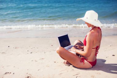 Gülümseyen kız bikini ve hasır şapka giyip denizde dizüstü bilgisayar kullanıyor.